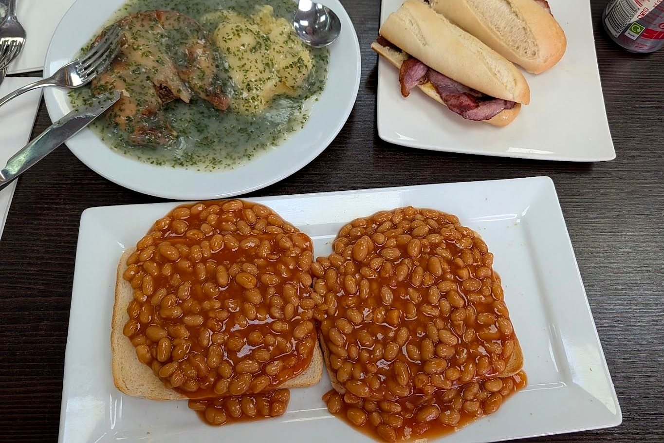 Beans on toast, a bacon baguette and a plate of food with green liquor on a dark café table
